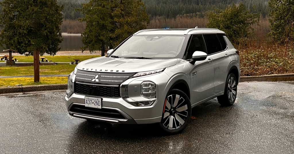 2026-Outlander-PHEV - Micro News - Calgary - Alberta - Canada Front-three-quarter view of a silver Mitsubishi Outlander SUV parked on a wet pavement with trees and a lake in the background, daytime outdoor scene.